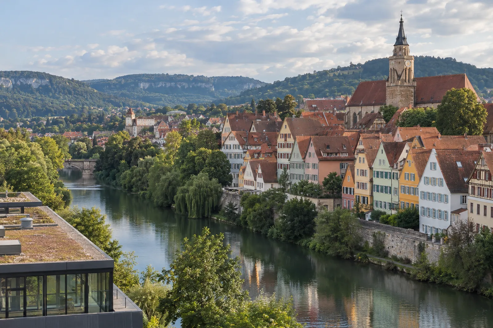 Neckarfront in Tübingen mit Altstadthäusern und Stiftskirche, im Vordergrund ein modernes Gewerbegebäude, dahinter die Schwäbische Alb