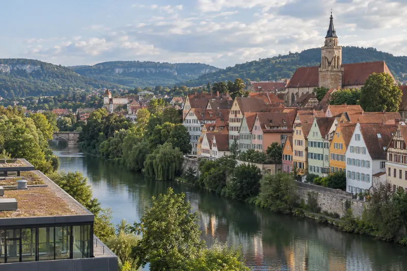 Neckarfront in Tübingen mit Altstadthäusern und Stiftskirche, im Vordergrund ein modernes Gewerbegebäude, dahinter die Schwäbische Alb