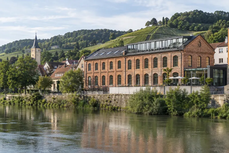 Ehemaliges Fabrikgebäude aus Backstein mit Rundbogenfenstern am Neckar, heute gewerblich genutzt, Weinberge im Hintergrund