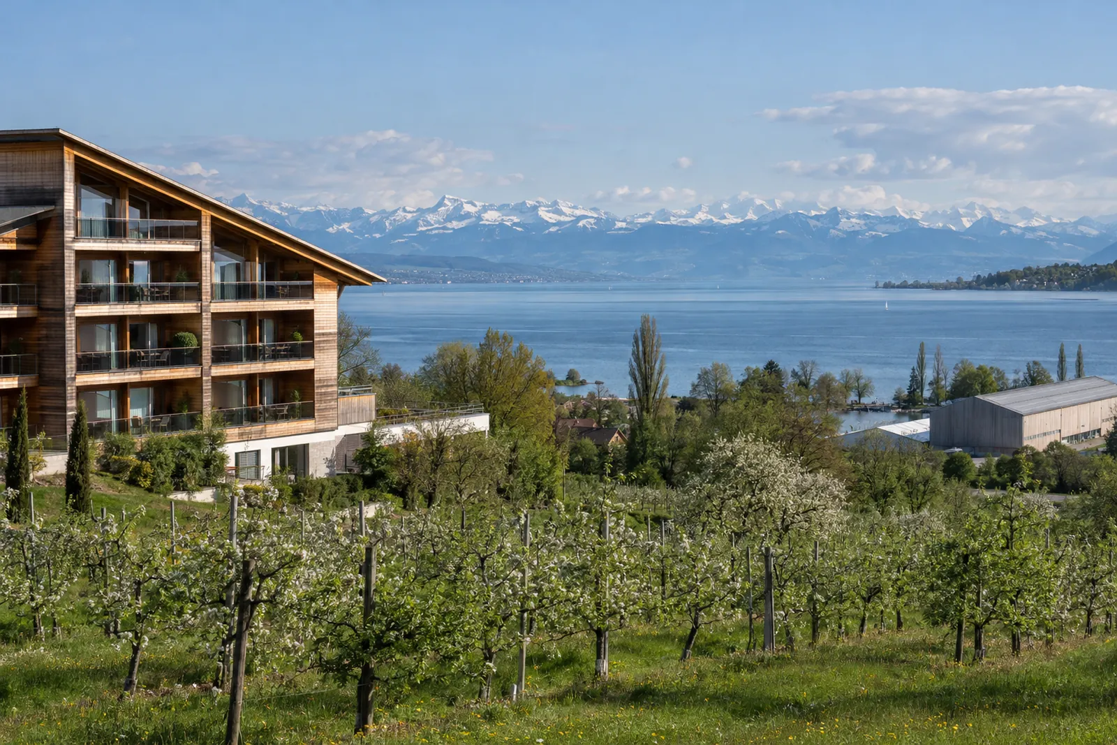 Hotelgebäude am Bodensee mit Holzbalkonen, im Hintergrund Wasser und Alpenkette
