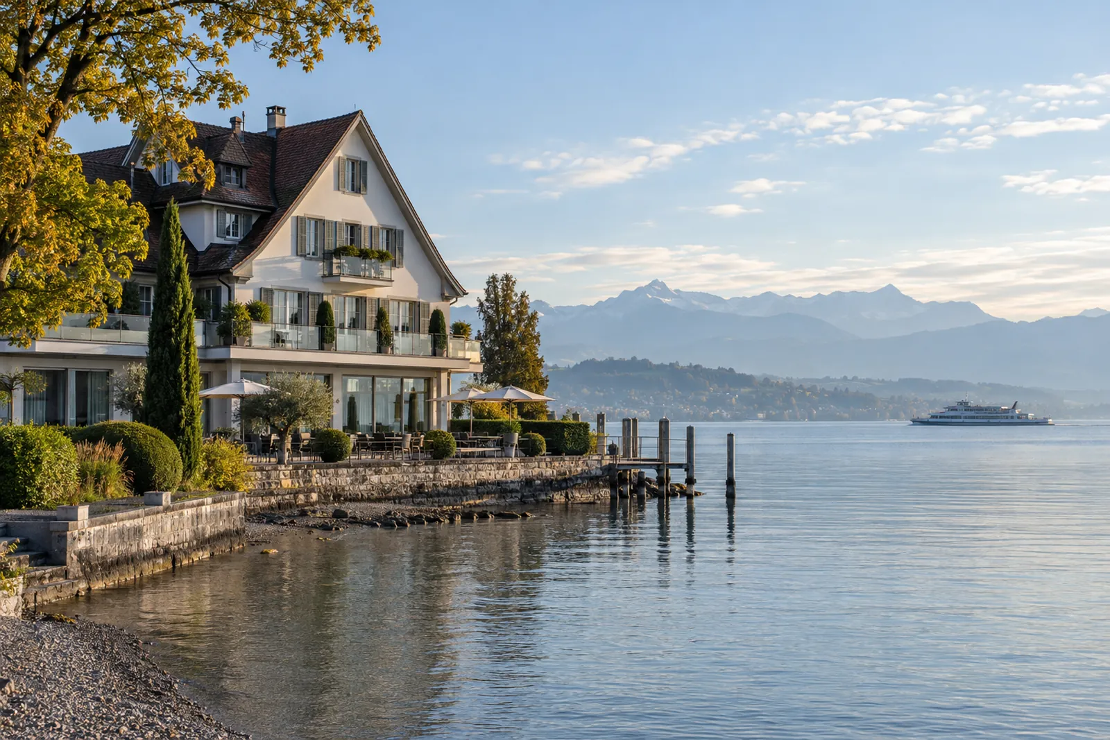 Kleines Hotel mit Satteldach am Bodenseeufer, im Hintergrund Alpenvorland