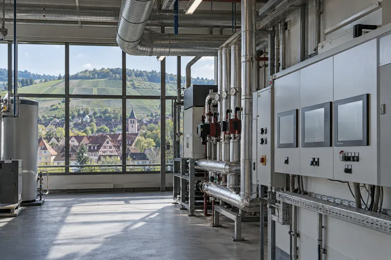 Technikraum einer Fertigungshalle mit Schaltschränken und Rohrleitungen, durch das Fenster Weinberge im Neckartal