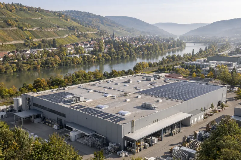 Produktionshalle mit Solardach im Neckartal, dahinter Weinberghänge und eine schwäbische Kleinstadt