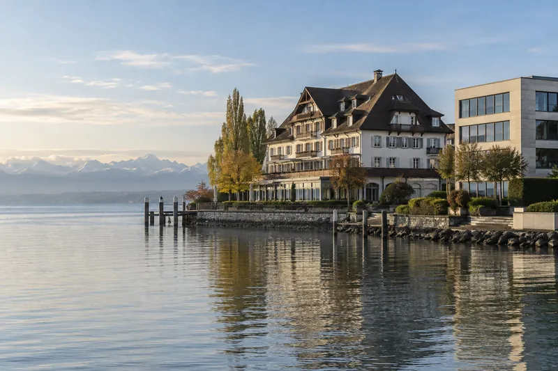 Hotel mit Satteldach am Bodensee, daneben ein schlichtes Bürogebäude, im Dunst die Alpen am gegenüberliegenden Ufer