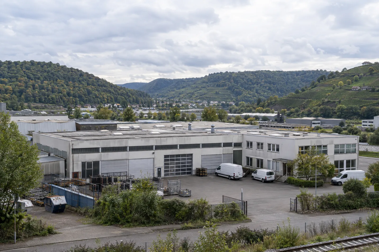 Ältere Produktionshalle mit Bürotrakt in einem Gewerbegebiet im Neckartal, dahinter bewaldete Hügel und Weinberge