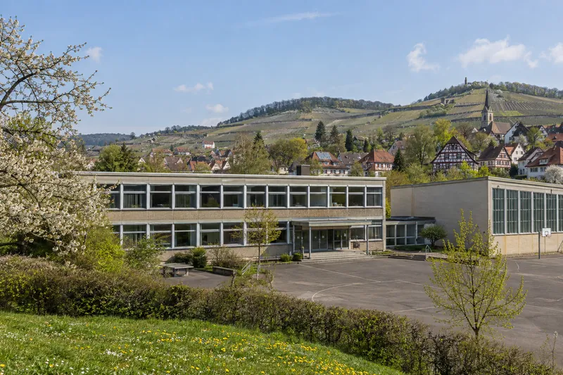 Schulgebäude der 1960er Jahre mit Flachdach und angebauter Turnhalle vor Weinbergen und Fachwerkhäusern in Baden-Württemberg