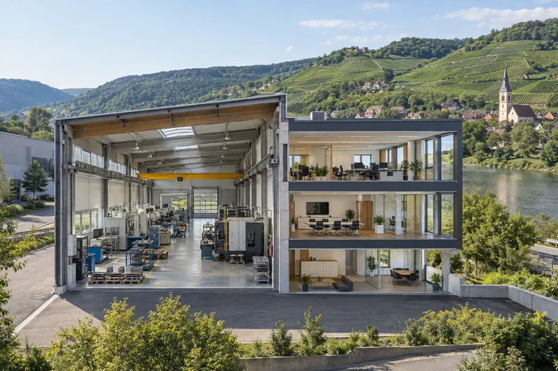 Gewerbebau im Neckartal mit Produktionshalle und Bürotrakt, im Hintergrund Weinberghänge und ein Kirchturm