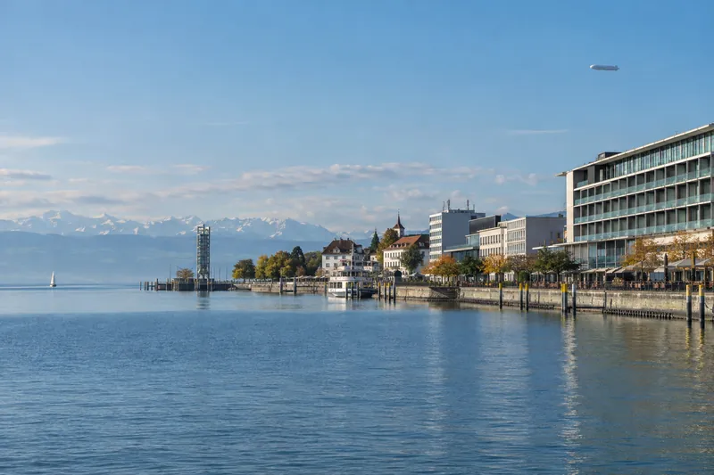 Hotel- und Geschäftsgebäude an der Hafenpromenade von Friedrichshafen mit Bodensee und Alpen im Hintergrund