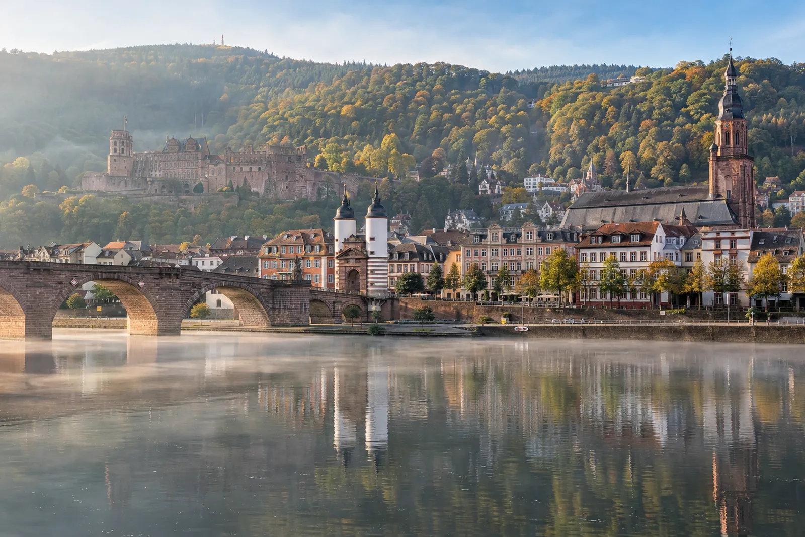 Geschäftshäuser und Hotels am Neckarufer in Heidelberg mit der Schlossruine am bewaldeten Hang im Hintergrund