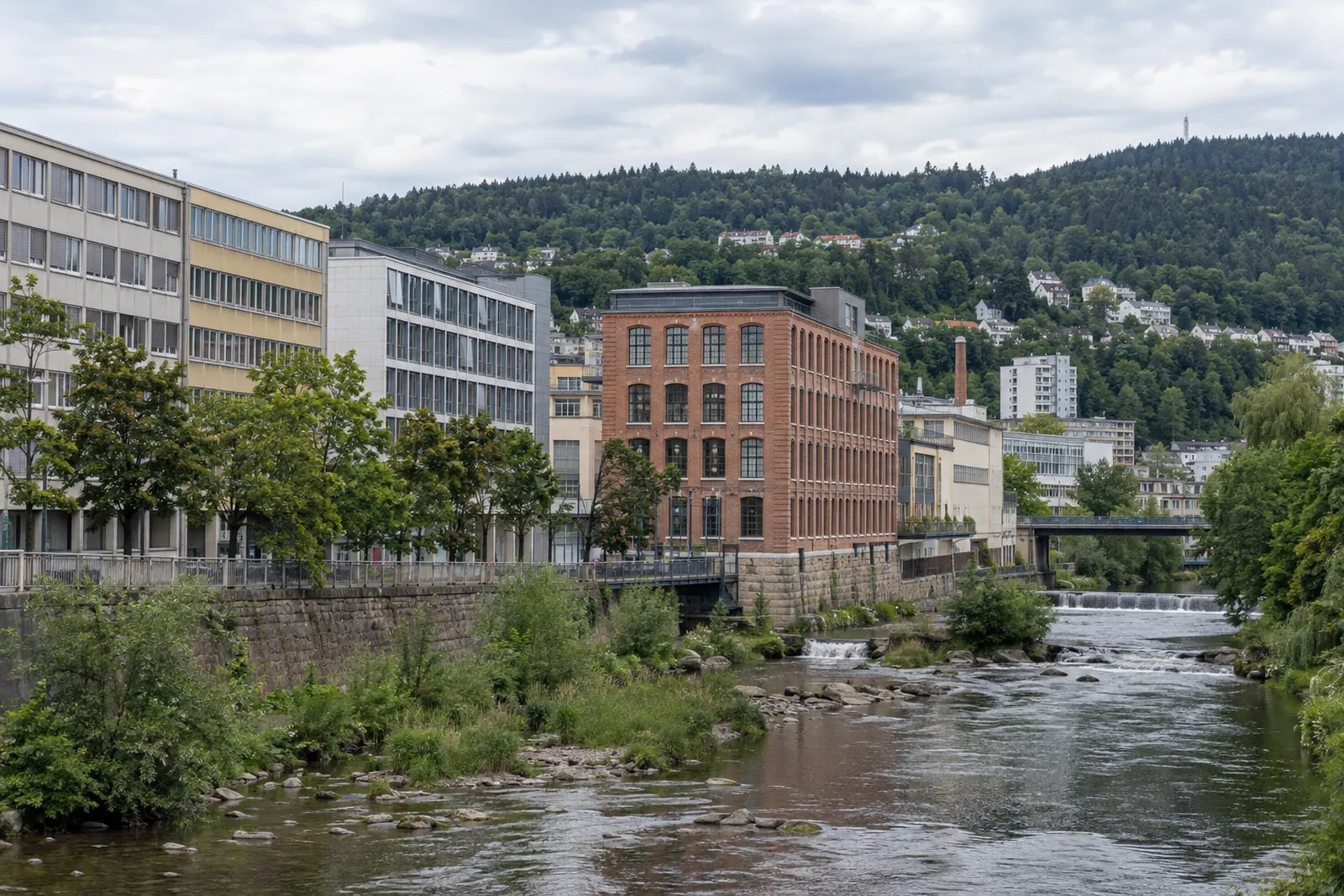 Gewerbebauten der Nachkriegszeit und ein Backsteinfabrikgebäude am Fluss in Pforzheim mit bewaldeten Höhen im Hintergrund