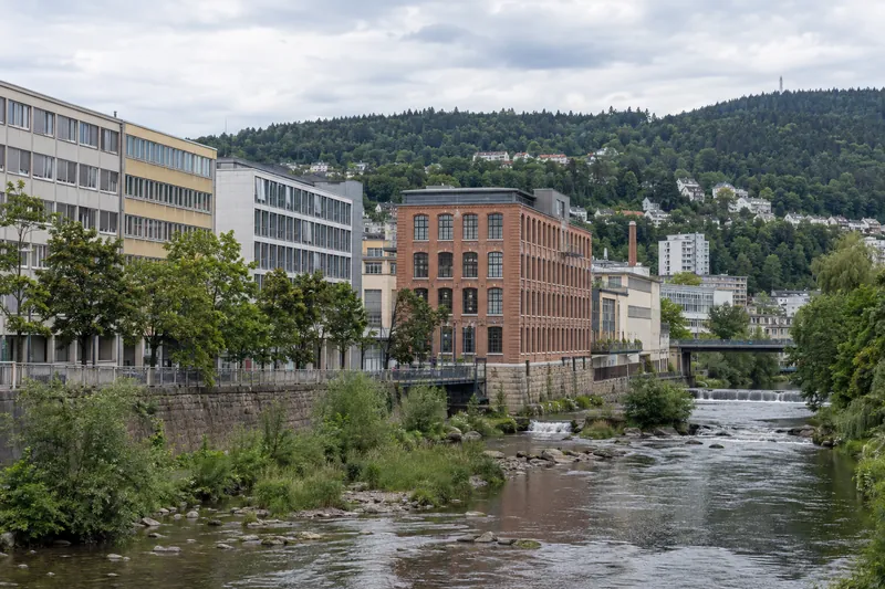 Gewerbebauten der Nachkriegszeit und ein Backsteinfabrikgebäude am Fluss in Pforzheim mit bewaldeten Höhen im Hintergrund